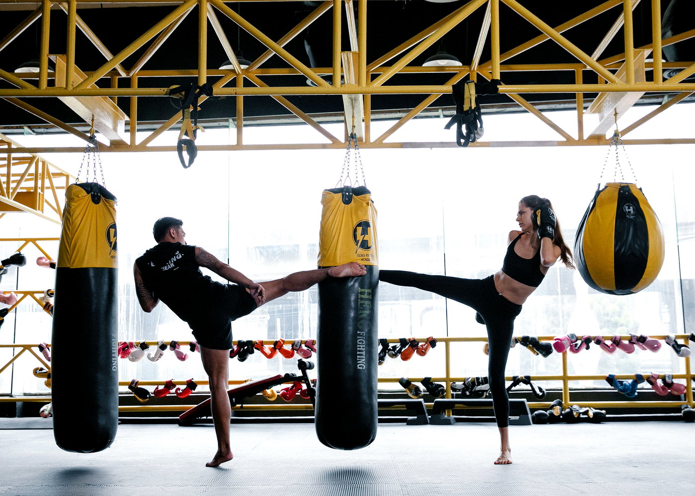 Two people kicking hanging punch bags in a gym with yellow steel beams and a rack of boxing gloves.