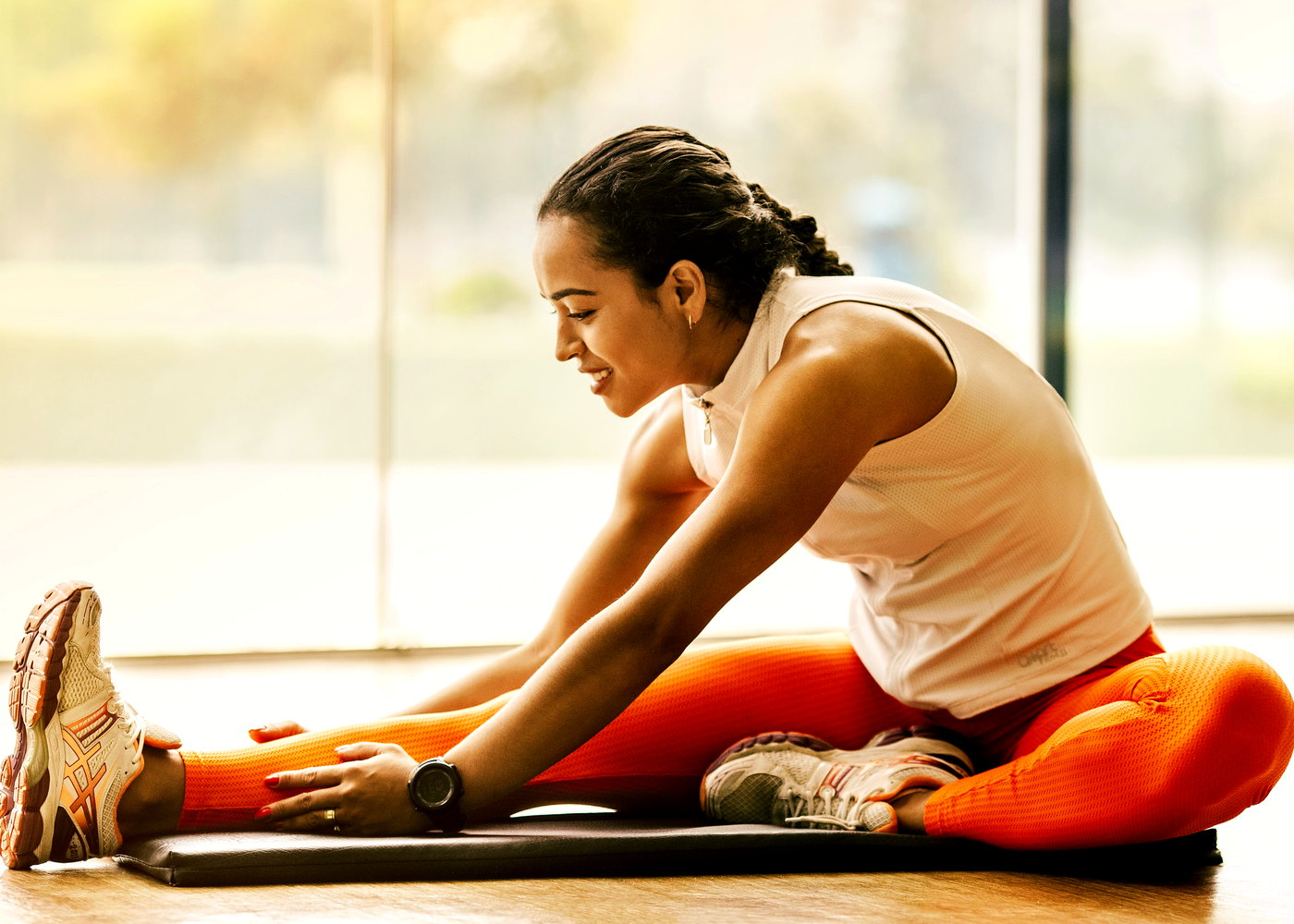 Woman stretching on a gym mat beside a window, wearing orange training tights.